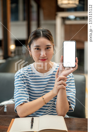 Asian young woman holding and showing blank screen smartphone while sitting at wooden table in cafe. Asian young woman holding and showing blank screen smartphone while sitting at wooden table in cafe. 135315112