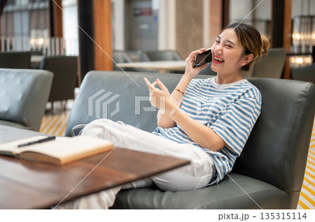 Asian young woman laughing during a phone call while sits on sofa with book on wooden table in cafe. 135315114