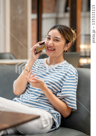 Asian young woman smiling during a phone call while sits on sofa with book on wooden table in a cafe. Asian young woman smiling during a phone call while sits on sofa with book on wooden table in a cafe. 135315115