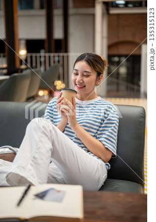Young asian woman looking at coffee holding with both hands while sitting leaning on sofa in a cafe. Young asian woman looking at coffee holding with both hands while sitting leaning on sofa in a cafe. 135315123