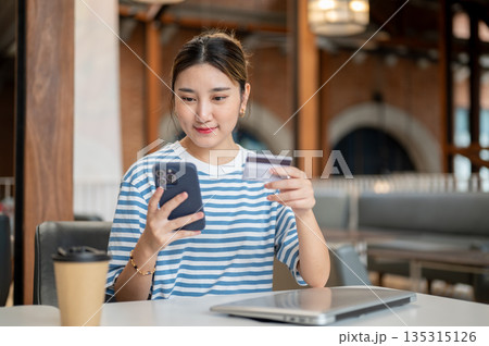 Asian woman holding credit card and looking at phone while sitting at table in cafe or coffee shop. Asian woman holding credit card and looking at phone while sitting at table in cafe or coffee shop. 135315126