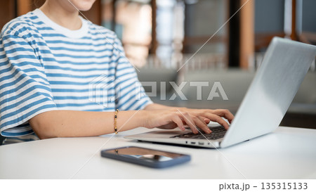 Close up of woman hands typing and working on laptop aside phone while sits at white table in a cafe Close up of woman hands typing and working on laptop aside phone while sits at white table in a cafe 135315133