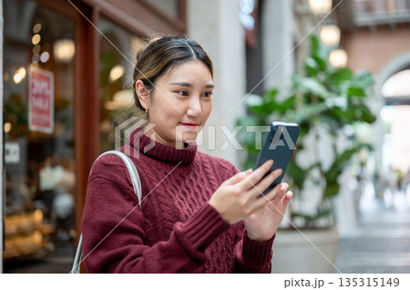 Young asian woman in knit sweater holding phone looking at picture taken in shopping mall or market. 135315149