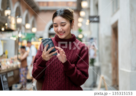 Smiling asian woman in knit sweater holding and looking at phone standing in shopping mall or market Smiling asian woman in knit sweater holding and looking at phone standing in shopping mall or market 135315153