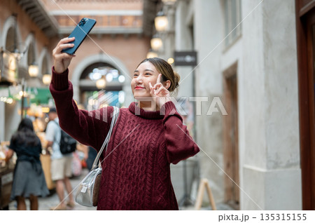 Asian woman in knit sweater holding phone taking picture doing peace sigh in shopping mall or market Asian woman in knit sweater holding phone taking picture doing peace sigh in shopping mall or market 135315155