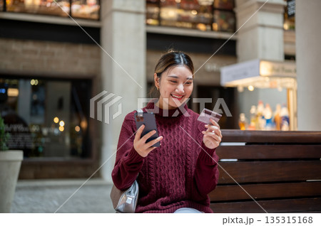 Asian woman holding credit card looking at phone while sitting on bench in shopping mall or market. 135315168