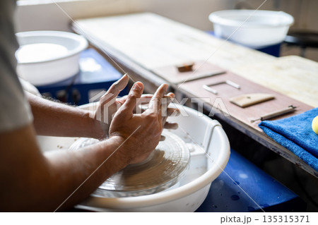 Close up of pottery artist's hands shaping a clay on spinning throwing wheel in a class or studio 135315371