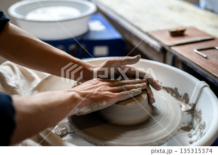Close up pottery artist hands making base of clay bowl on spinning throwing wheel in class or studio Close up pottery artist hands making base of clay bowl on spinning throwing wheel in class or studio 135315374