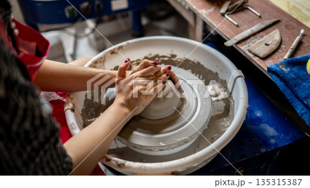 Woman pottery artist with painted nails molding a clay on spinning throwing wheel in class or studio 135315387