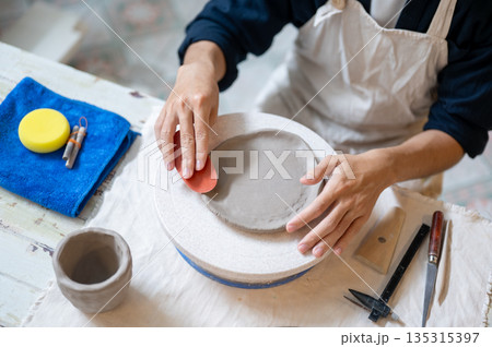 Pottery artist using a scraper smoothing a clay on throwing wheel at working table in class or studio. Pottery artist using a scraper smoothing a clay on throwing wheel at working table in class or studio. 135315397