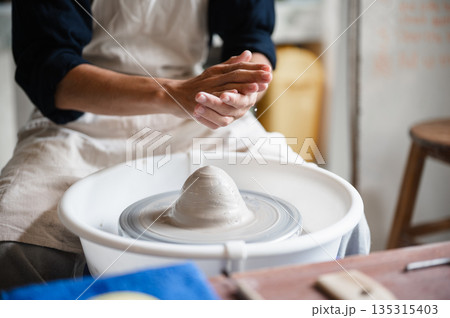 Close up of pottery artist's hands preparing to mold a clay on throwing wheel in a class or studio. Close up of pottery artist's hands preparing to mold a clay on throwing wheel in a class or studio. 135315403