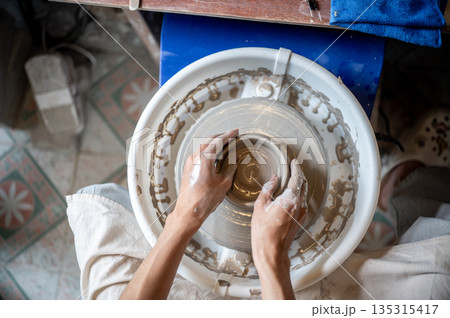 Close up of pottery artist's hands gripping edge of clay bowl on throwing wheel in class or studio. Close up of pottery artist's hands gripping edge of clay bowl on throwing wheel in class or studio. 135315417