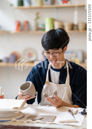 Young man potter artist holding handle and clay mug on working table in a pottery class or studio. 135315509