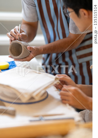 Pottery teacher holding brush showing how to add texture on clay mug on table in class or studio. Pottery teacher holding brush showing how to add texture on clay mug on table in class or studio. 135315514