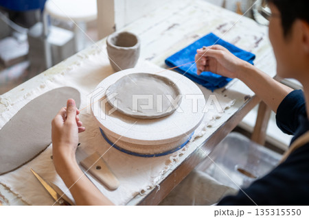 Pottery artist using wire cutter on clay slab on throwing wheel at working table in class or studio Pottery artist using wire cutter on clay slab on throwing wheel at working table in class or studio 135315550