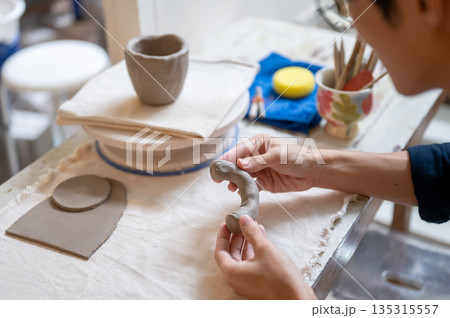 Close up of pottery artist's hand molding clay into a mug handle at working table in class or studio Close up of pottery artist's hand molding clay into a mug handle at working table in class or studio 135315557
