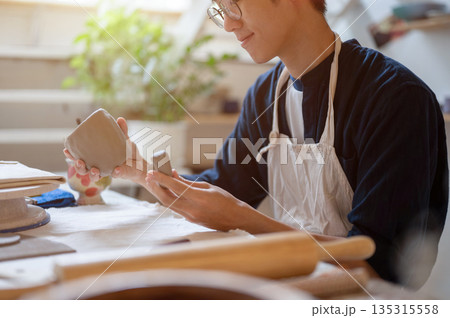Young pottery artist holding and looking at clay mug with handle at working table in class or studio Young pottery artist holding and looking at clay mug with handle at working table in class or studio 135315558