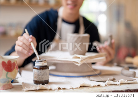 Potter artist holding brush over slip bottle for gluing clay mug at working table in class or studio 135315571