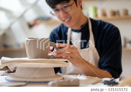 Pottery artist holding handle add to clay mug on throwing wheel at working table in a class or studio 135315572