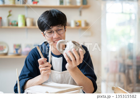 Young pottery artist holding and looking at a complete clay mug at working table in class or studio. Young pottery artist holding and looking at a complete clay mug at working table in class or studio. 135315582
