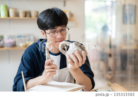 Glasses pottery artist holding and looking at complete clay mug at working table in class or studio Glasses pottery artist holding and looking at complete clay mug at working table in class or studio 135315583