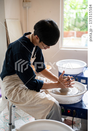 Young pottery artist's with stained hands shaping up a clay on throwing wheel in a class or studio. 135315584