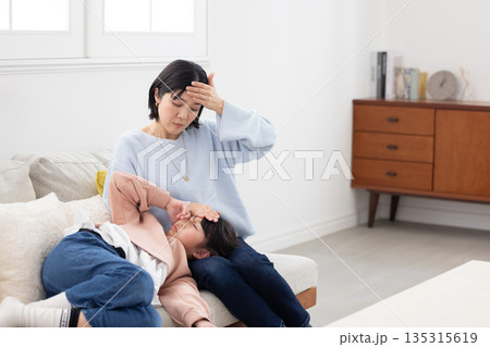 膝の上に寝かせた娘の額に手を当てて熱を計る母親 膝の上に寝かせた娘の額に手を当てて熱を計る母親 135315619