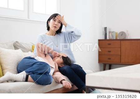 膝の上に寝かせた娘の額に手を当てて熱を計る母親 膝の上に寝かせた娘の額に手を当てて熱を計る母親 135315620