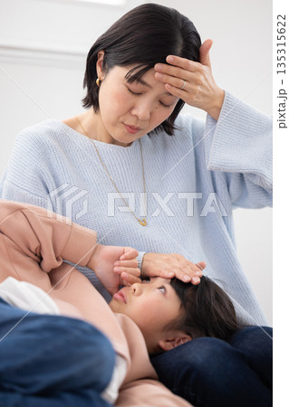 膝の上に寝かせた娘の額に手を当てて熱を計る母親 膝の上に寝かせた娘の額に手を当てて熱を計る母親 135315622