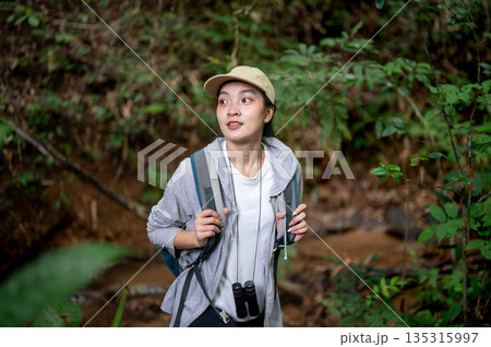 Asian woman tourist walking holding bag straps and looking around during forest hiking or trekking. Asian woman tourist walking holding bag straps and looking around during forest hiking or trekking. 135315997