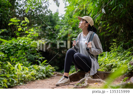 Woman backpacker holding bag strap sitting on ground tree root during solo forest hiking or trekking Woman backpacker holding bag strap sitting on ground tree root during solo forest hiking or trekking 135315999