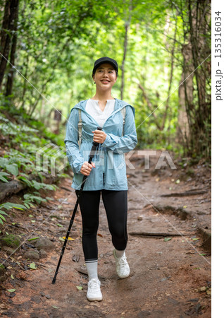 Pretty asian tourist woman looking ahead walking downhill holding trekking cane as hiking in forest. Pretty asian tourist woman looking ahead walking downhill holding trekking cane as hiking in forest. 135316034