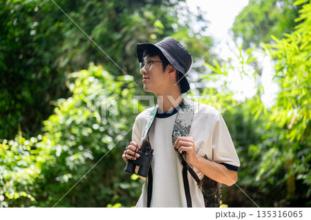 Backpacker tourist man holding binoculars looking around while solo hiking or trekking in the forest 135316065