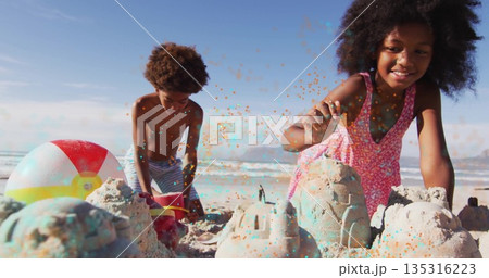 Siblings in pink dress and trunks building sandcastles on beach with red bucket, plastic shovel Siblings in pink dress and trunks building sandcastles on beach with red bucket, plastic shovel 135316223