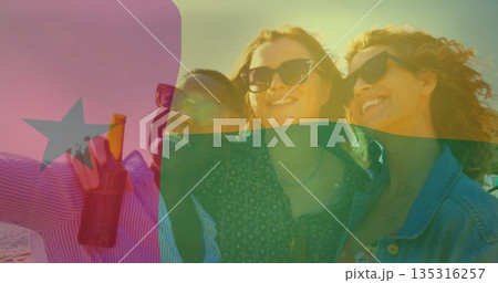 Smiling three friends posing at beach at sunset, with bottle and sunglasses, Ghanaian flag overlay 135316257
