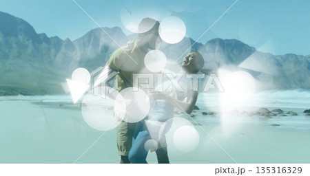 Romantic couple dipping and laughing on sunlit wet sand at beach shore, mountain peaks 135316329