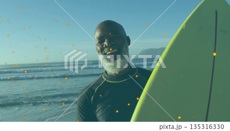 Standing bearded man holding green-yellow surfboard at ocean edge, wearing black rash guard 135316330
