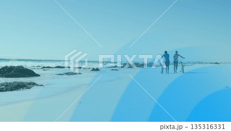 Walking couple in sportswear pushing bicycles along beach with rocks and lapping waves, copy space 135316331