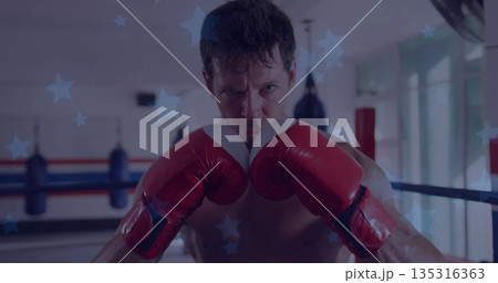 Holding red boxing gloves, shirtless boxer defending in gym ring, with ring ropes and punching bags 135316363