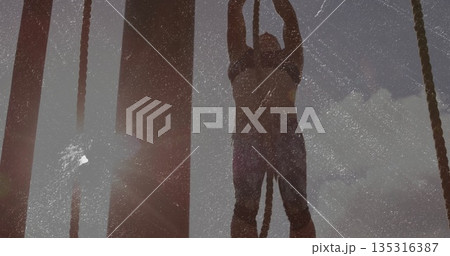 Climbing woman gripping thick rope and wooden posts on obstacle course, in sports bra and leggings 135316387