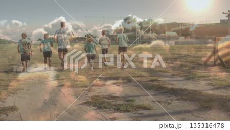 Jogging coach and children wearing shorts and t-shirts across sports field, with soccer goal frames Jogging coach and children wearing shorts and t-shirts across sports field, with soccer goal frames 135316478
