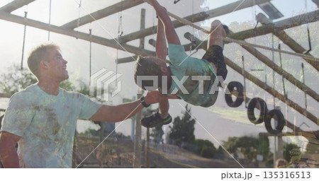 Steadying parent wearing sportswear supporting child hanging on monkey bars in park, hanging tires 135316513