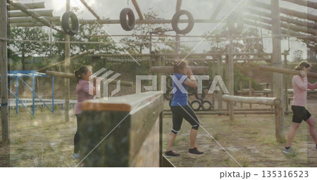 Walking group of five women in activewear carrying logs at obstacle course, with hanging tires 135316523