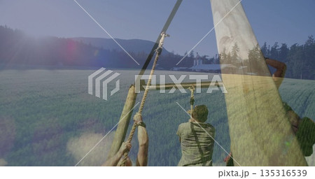 Climbing obstacle frame in green field wearing gloves and straps gripping thick ropes, barn silos Climbing obstacle frame in green field wearing gloves and straps gripping thick ropes, barn silos 135316539