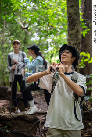 Tourist man holding binoculars standing looking up in front of friends in forest hiking or trekking Tourist man holding binoculars standing looking up in front of friends in forest hiking or trekking 135316642