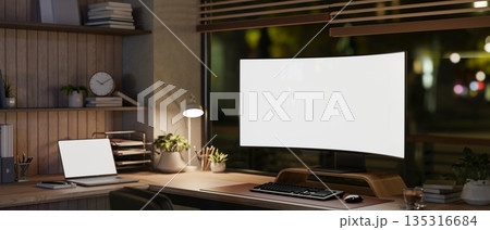 Blank curved screen computer and laptop under lamp light on wooden table in dim lit room or office. 135316684