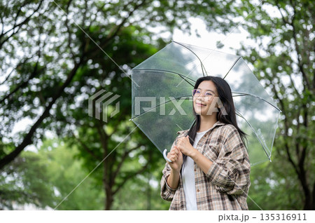 Young asian glasses woman smiling as standing holding transparent umbrella in rain at park or garden Young asian glasses woman smiling as standing holding transparent umbrella in rain at park or garden 135316911