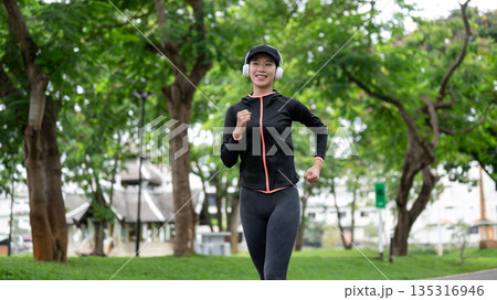 Young asian woman in sportswear with headphone on listening to music as exercise by running in park. Young asian woman in sportswear with headphone on listening to music as exercise by running in park. 135316946