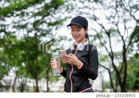 Young asian woman in sportswear holding water and looking at phone after exercise or workout in the park. Young asian woman in sportswear holding water and looking at phone after exercise or workout in the park. 135316955
