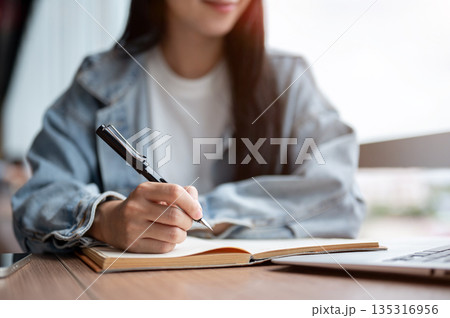 Close up of woman in denim jacket holding fountain pen writing in notebook on wooden table in cafe. Close up of woman in denim jacket holding fountain pen writing in notebook on wooden table in cafe. 135316956